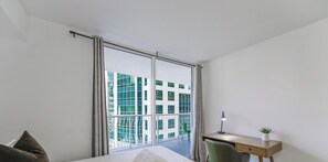 Quarto com uma janela grande, uma escrivaninha, uma cadeira e uma cama.
