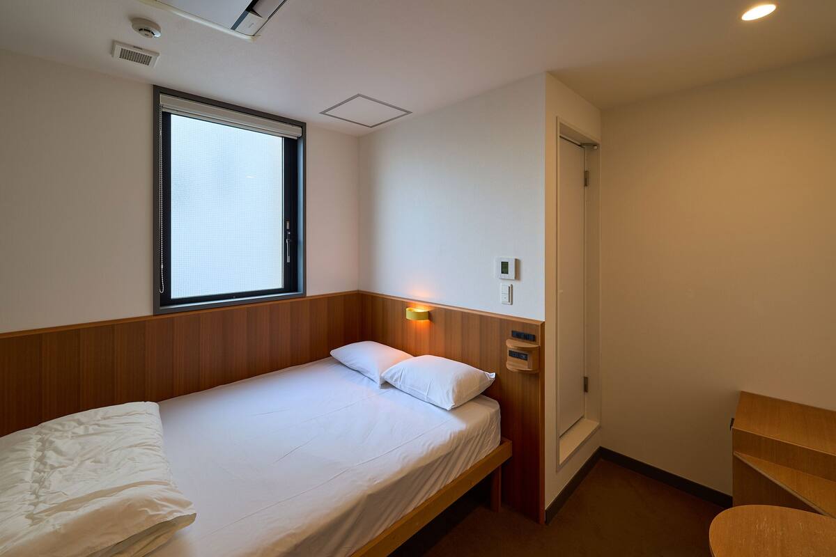 A single bed with a wooden headboard, a bedside table, a wall-mounted light, and a window with blinds.