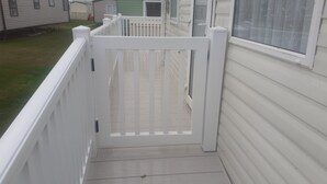 A white wooden deck with a white railing, leading to a house with a green door and a window with a decorative screen. - Pet friendly 2 bed caravan in coopers beach (Mersea Island)