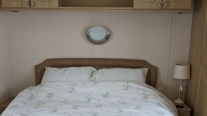 A bedroom with a wooden headboard, a round mirror above the bed, and a nightstand with a lamp. - Pet friendly 2 bed caravan in coopers beach (Mersea Island)