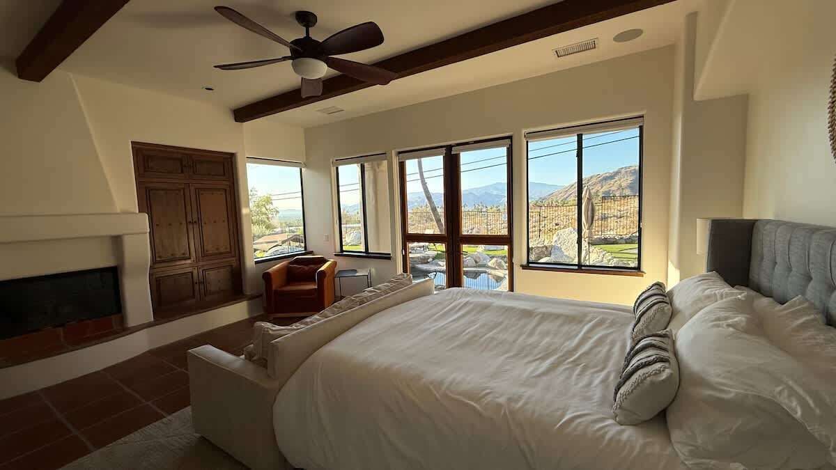 A bedroom with a large bed, a fireplace, a ceiling fan, and a view of mountains through large windows.