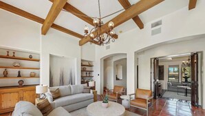 Ein geräumiges Wohnzimmer mit hohen Decken, Holzbalken, einem Kronleuchter, einer Sofagarnitur, einem Couchtisch und Blick in den Essbereich.