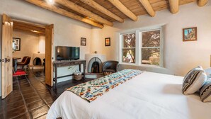 Een slaapkamer met een groot bed, houten balken in het plafond, een open haard, een televisietoestel en een raam met uitzicht op bomen.