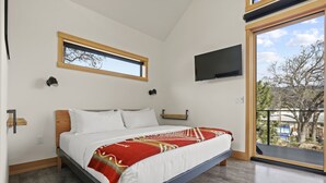 A modern bedroom with a large bed, a TV mounted on the wall, a window with a view of trees, and a balcony.