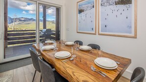A dining area with a wooden table set for six, large windows with a mountain view, and two framed pictures on the wall. - Saddle Ridge Ski in/Ski out condo, private hot tub (Big Sky)