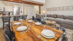A dining area with a wooden table set for four, surrounded by chairs. A sofa and a coffee table are in the background. - Saddle Ridge Ski in/Ski out condo, private hot tub (Big Sky)