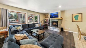 A living room with a sofa, coffee table, fireplace, TV, and large windows. - Family Cabin with Hot Tub and Game Room (South Lake Tahoe)