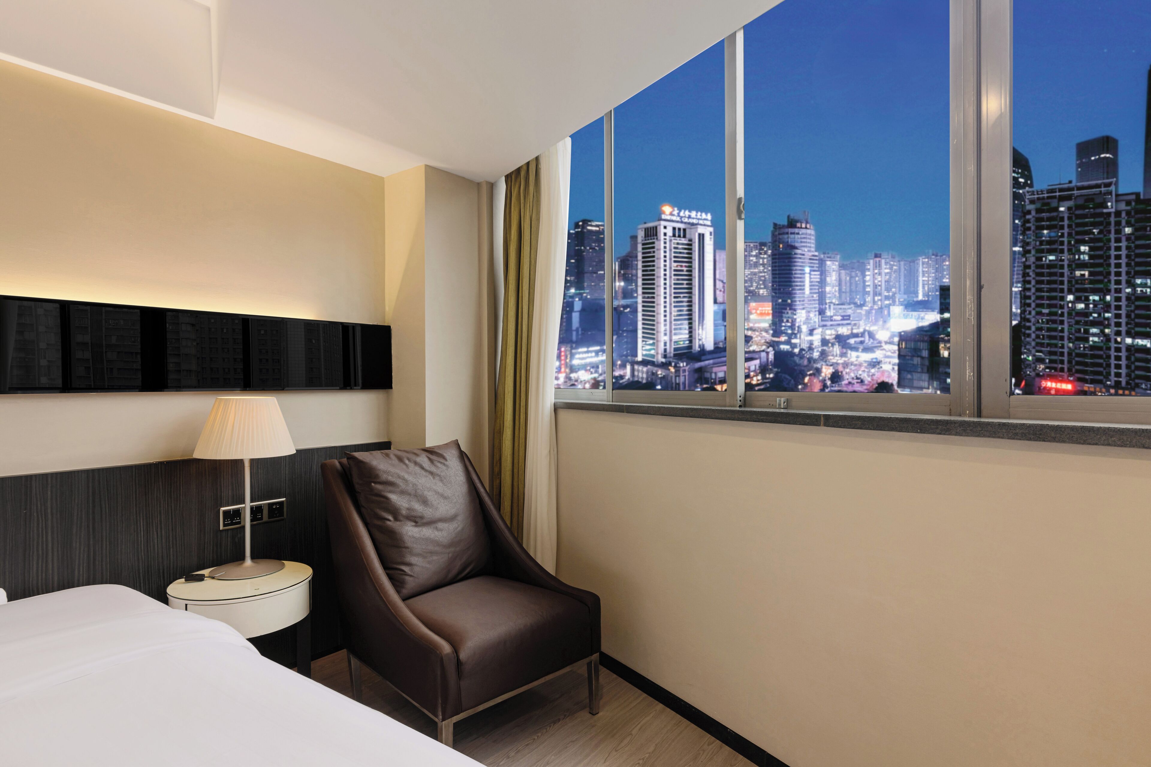 Habitación de hotel con un ventanal amplio que ofrece vistas nocturnas de la ciudad, una cama, una silla y una mesita de noche con lámpara.