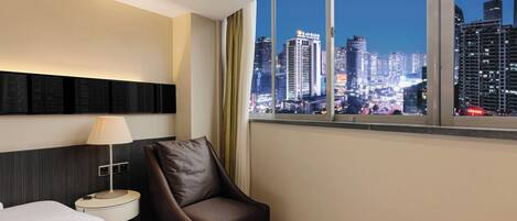 Ein Hotelzimmer mit großem Fenster, das einen Blick auf die nächtliche Stadt bietet, ein Bett, ein Sessel und ein Nachttisch mit Lampe.