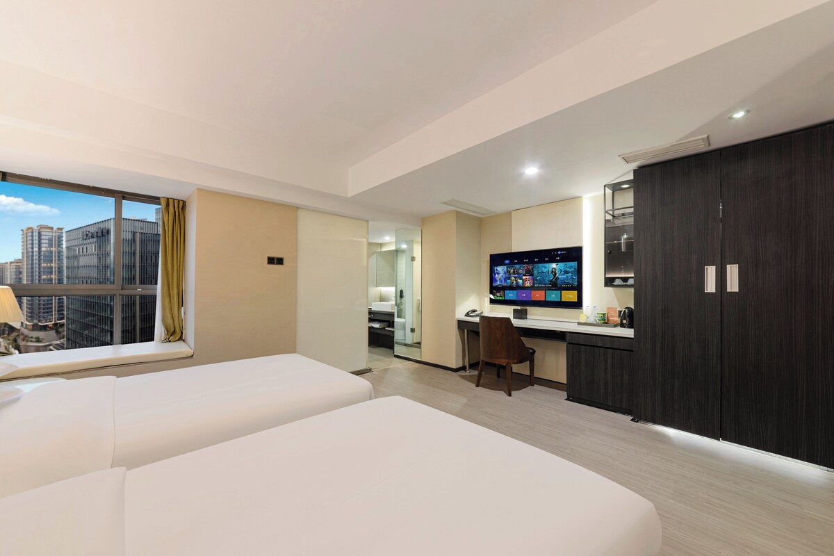 A hotel room with two beds, a desk, a TV, and a view of the cityscape.