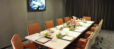 A conference room with a long table, orange chairs, a TV on the wall, and a floral centerpiece.