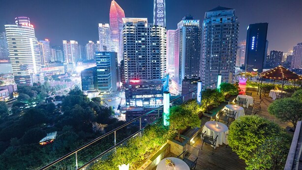 Vista urbana noturna com edifícios iluminados e um jardim no terraço com mesas e cadeiras.