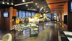 Uma sala de jantar moderna com mesas redondas, estações de buffet e uma grande janela com vista para a cidade.