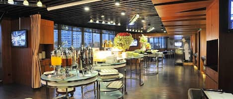 A modern dining area with round tables, buffet stations, and a large window providing a city view.