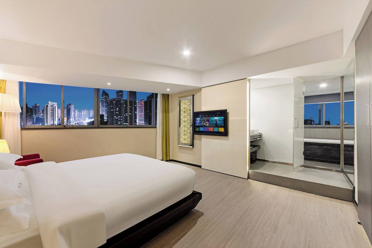 A modern hotel room with a large bed, a TV mounted on the wall, and a city view through large windows.