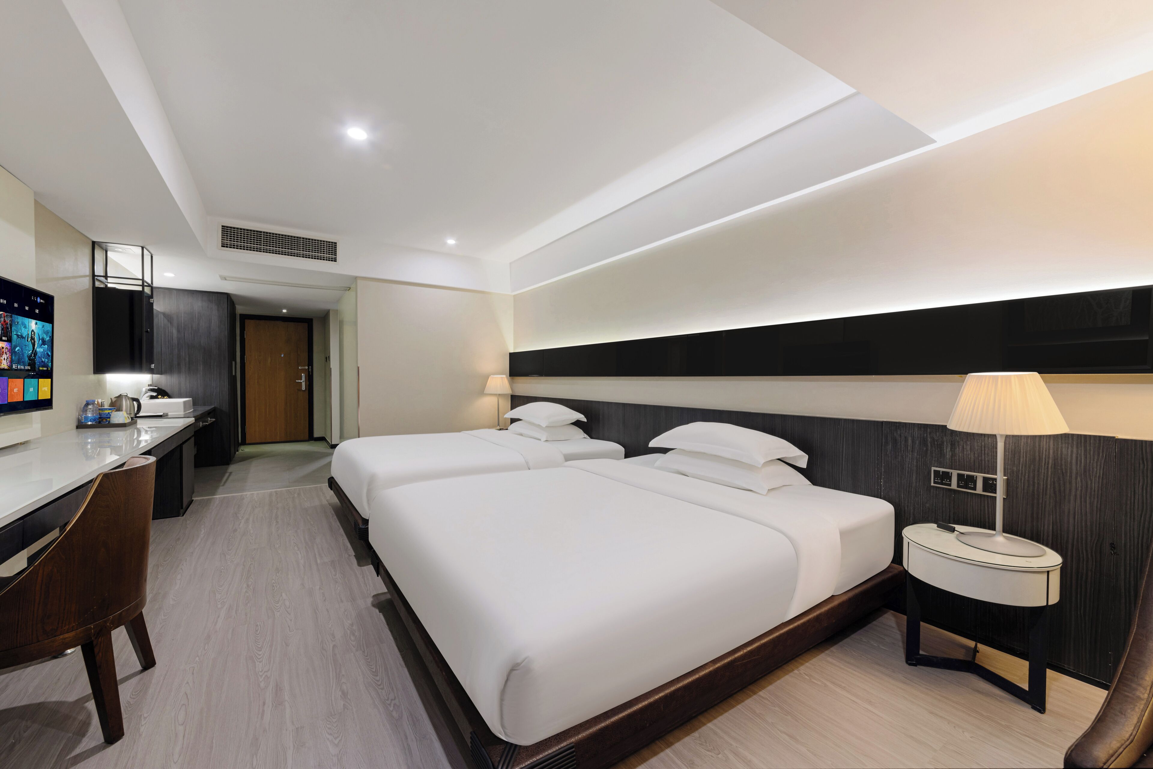 A hotel room with two beds, a desk, a chair, a TV, and a lamp.