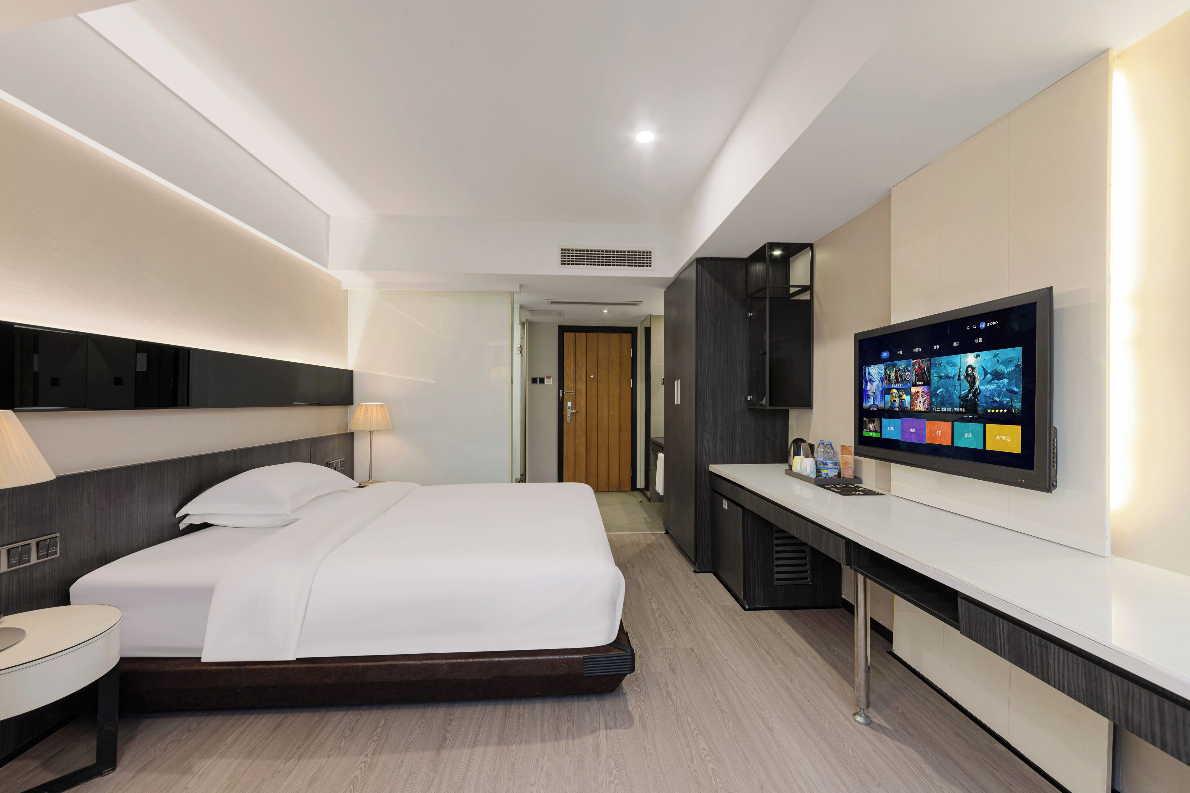 Ein modernes Hotelzimmer mit Bett, Nachttisch, Lampe, Fernseher und einer kleinen Küchenzeile.