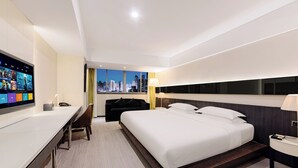 Quarto de hotel moderno com uma cama grande, uma mesa com televisão de tela plana, vista para a cidade e mesa de cabeceira com abajur.