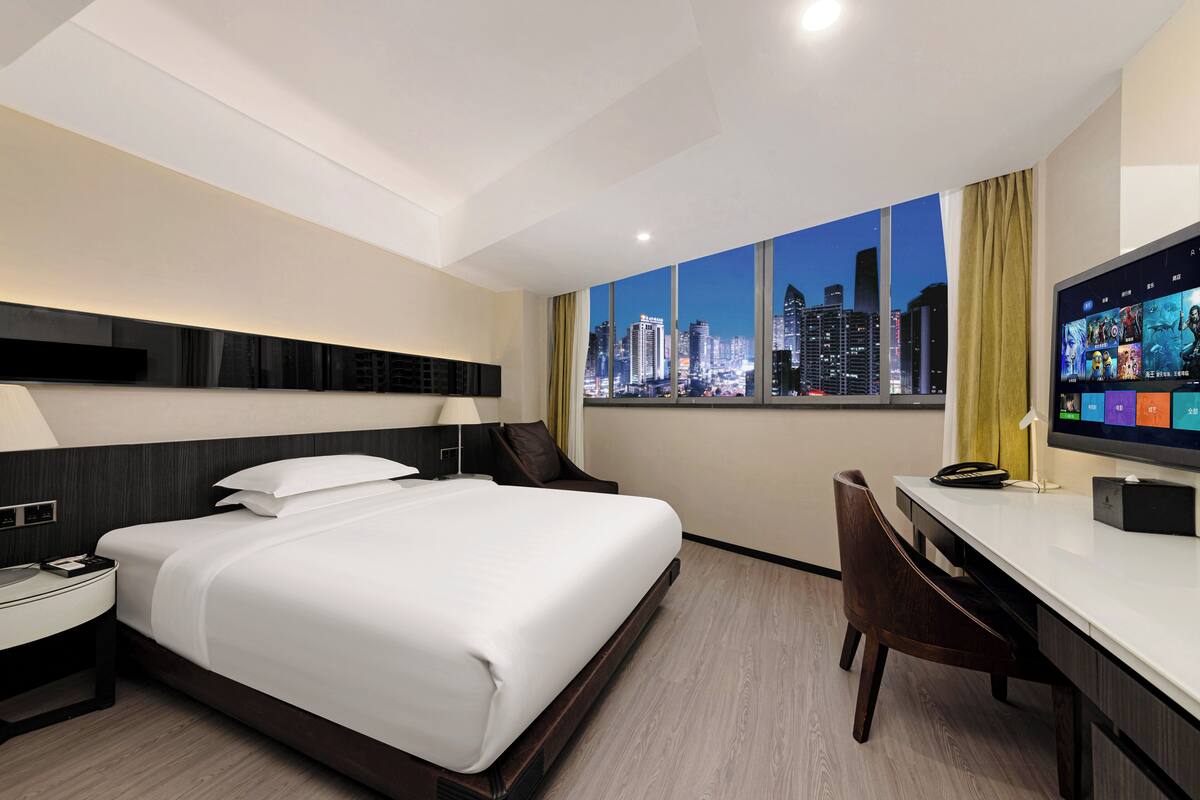 A hotel room with a bed, desk, chair, and a large window with a cityscape view.