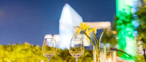 A table with two wine glasses and a candle, set against a backdrop of greenery and a building.