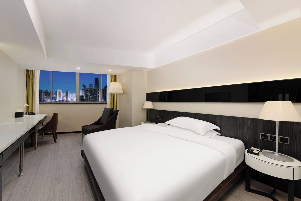 A hotel room with a large bed, a desk, a chair, a nightstand with a lamp, and a view of a city skyline at dusk.