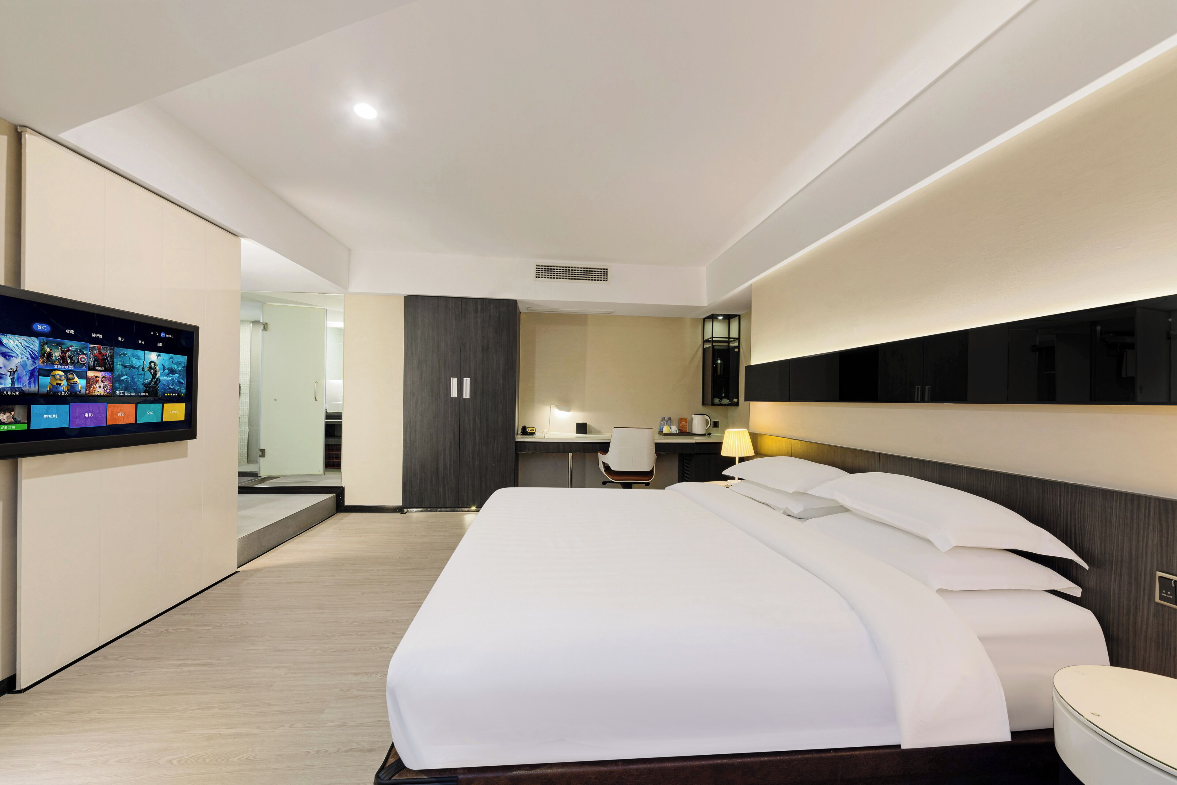Quarto de hotel moderno com uma cama grande, escrivaninha, poltrona, televisão fixada na parede e abajur.