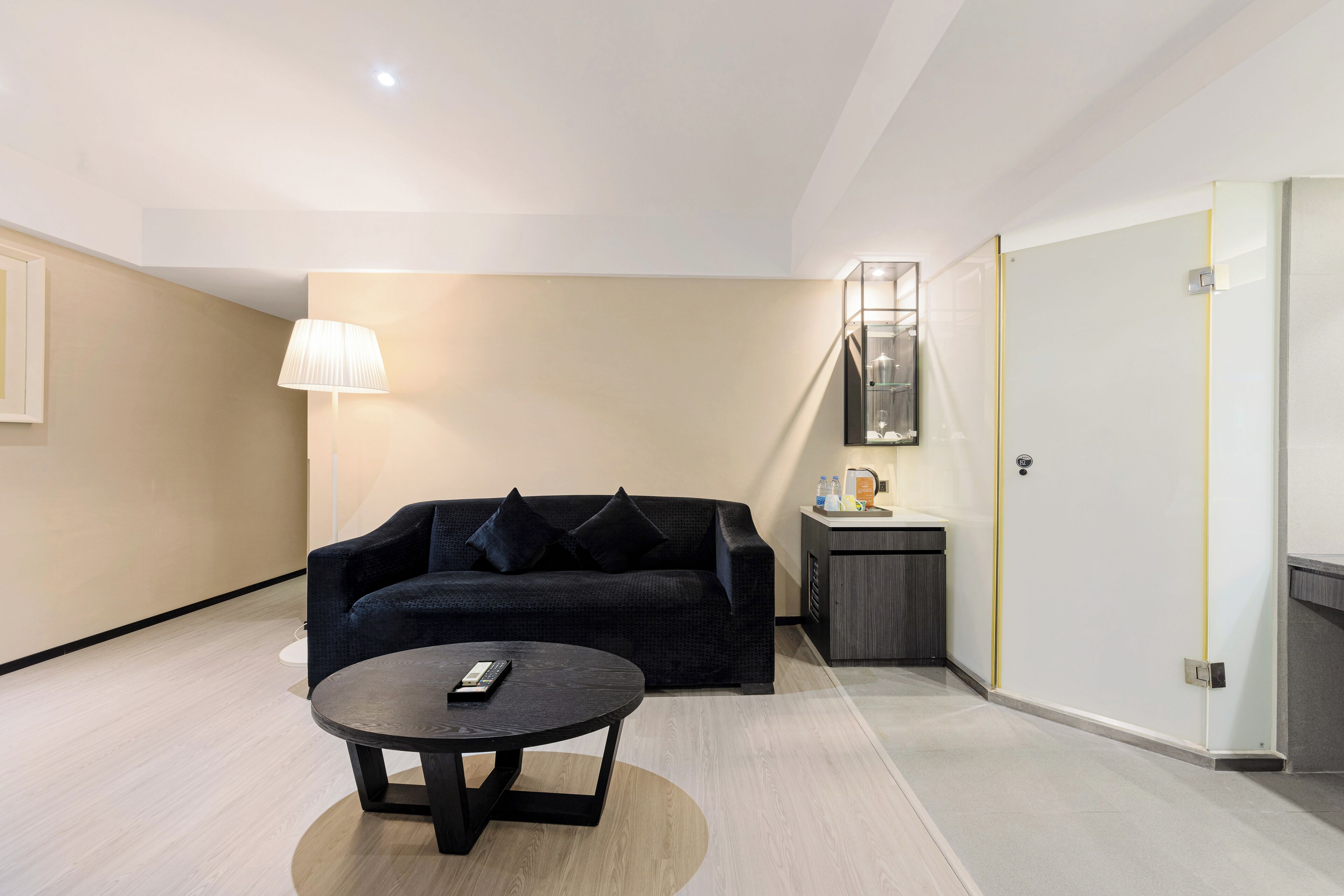 A modern living room with a dark sofa, a round coffee table, a lamp, and a small kitchenette area.