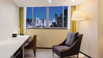 Habitación de hotel con escritorio, silla y ventana grande que ofrece vista panorámica de la ciudad.