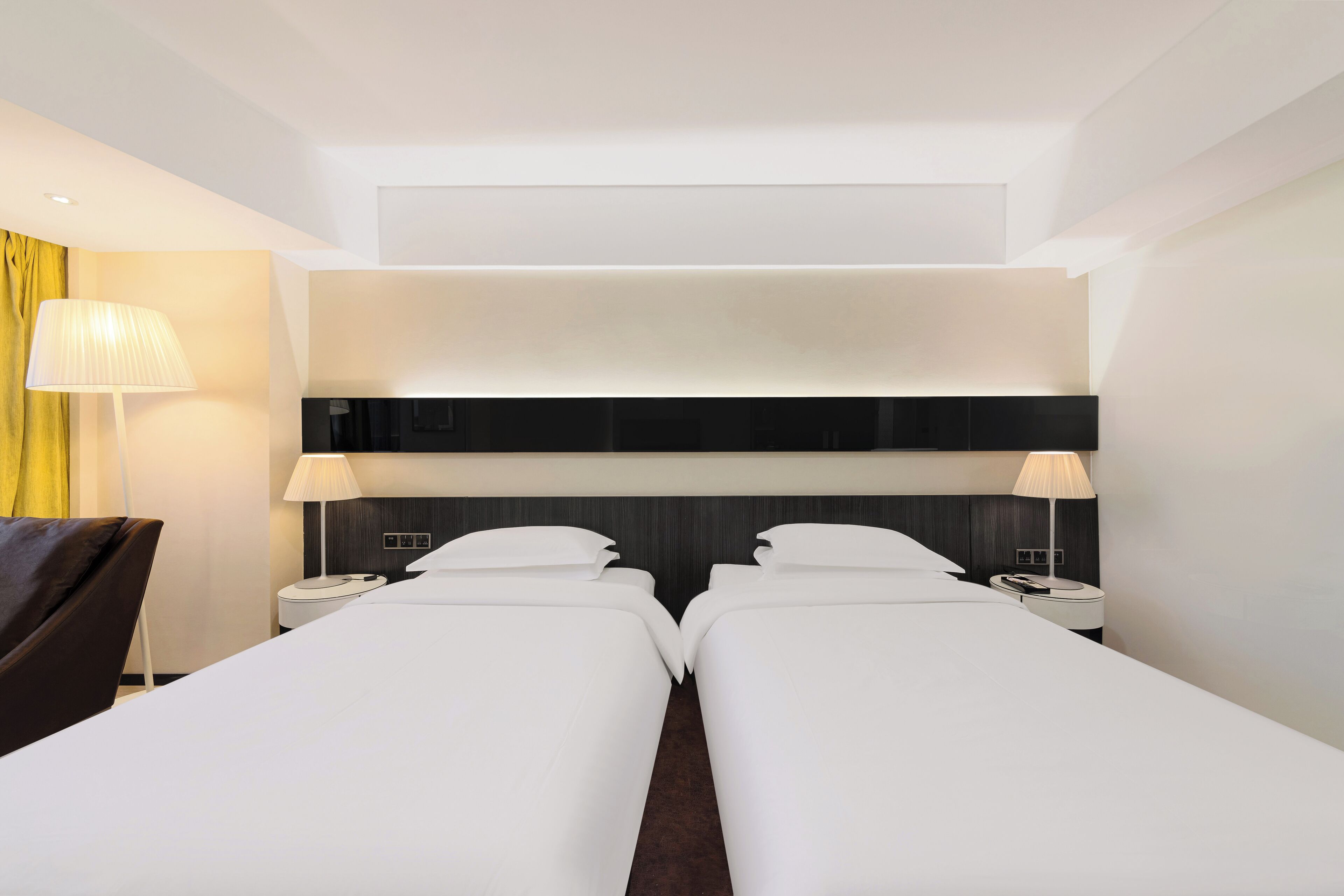 A hotel room with two single beds, a dark headboard, bedside lamps, and a yellow curtain.