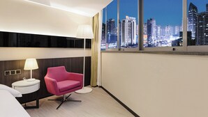 Habitación de hotel moderna con un amplio ventanal que ofrece vistas a la ciudad, una mesita redonda con lámpara y una silla rosa.