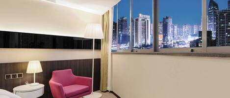 A modern hotel room with a large window offering a city view, a round side table with a lamp, and a pink chair.