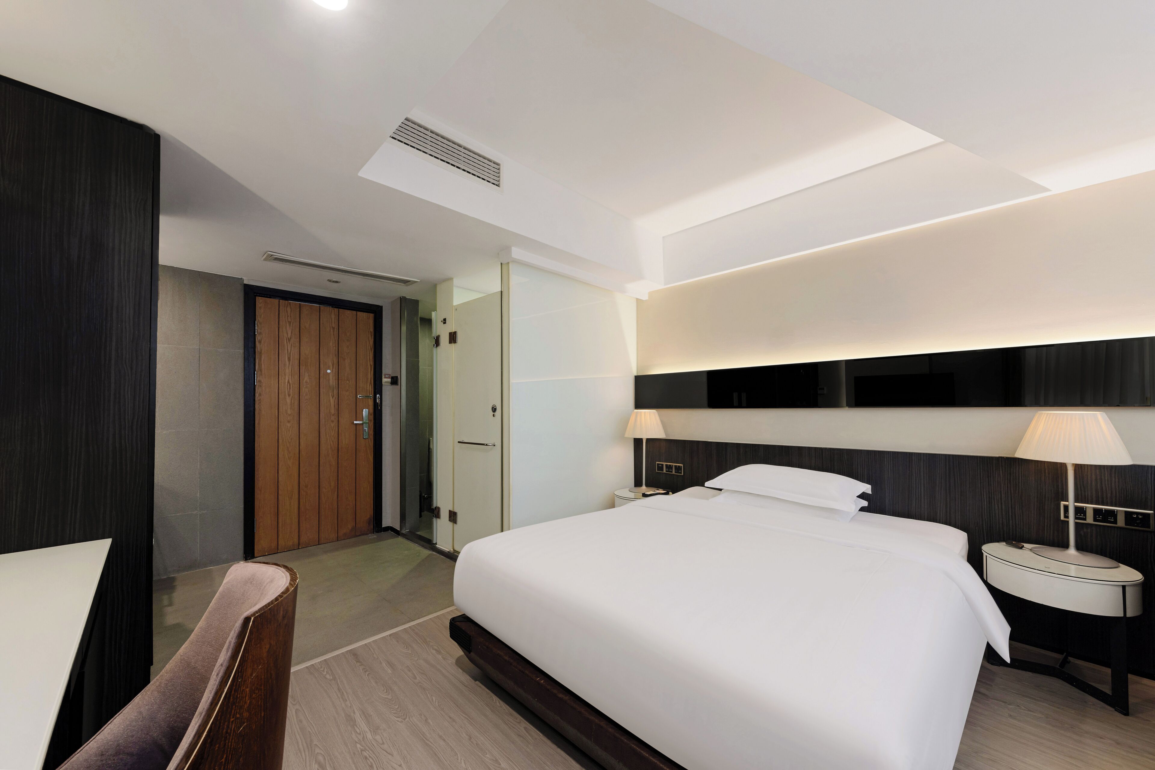 A modern hotel room with a large bed, bedside tables with lamps, a wooden door, and a ceiling vent.