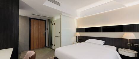 A modern hotel room with a large bed, bedside tables with lamps, a wooden door, and a ceiling vent.