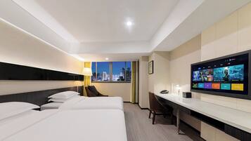 Habitación de hotel con tres camas, un escritorio, televisión y vista a la ciudad.
