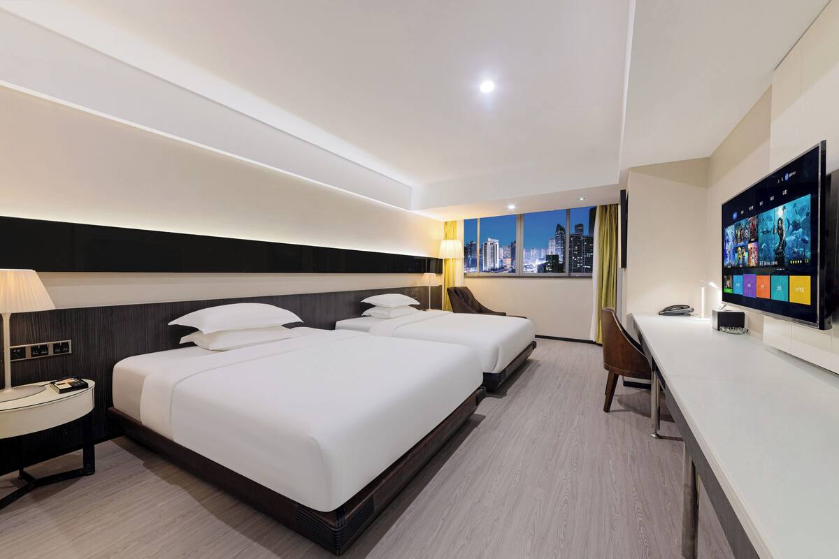A modern hotel room with two beds, a desk, a TV, and a city view.
