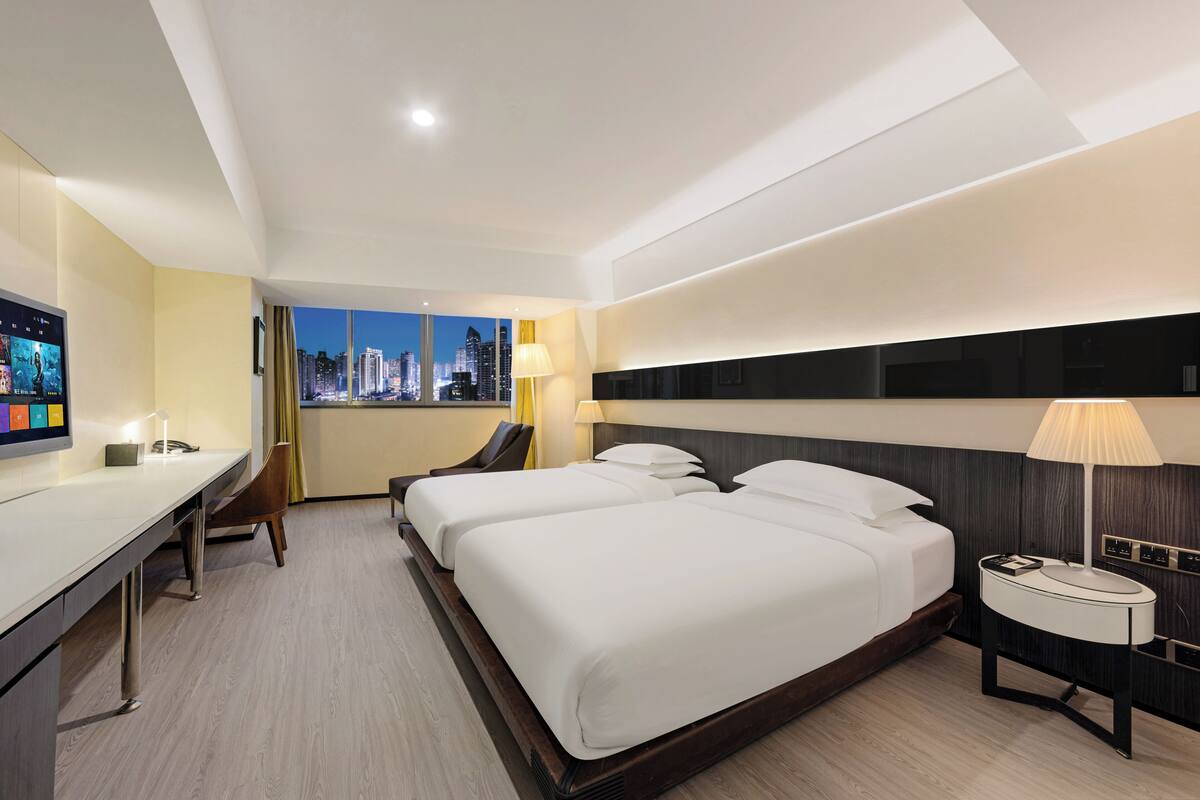 A hotel room with two beds, a desk, a chair, a nightstand, and a view of a cityscape at night.