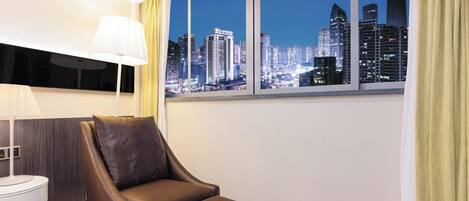A modern hotel room with a cityscape view, a leather armchair, a bedside table, and a lamp.