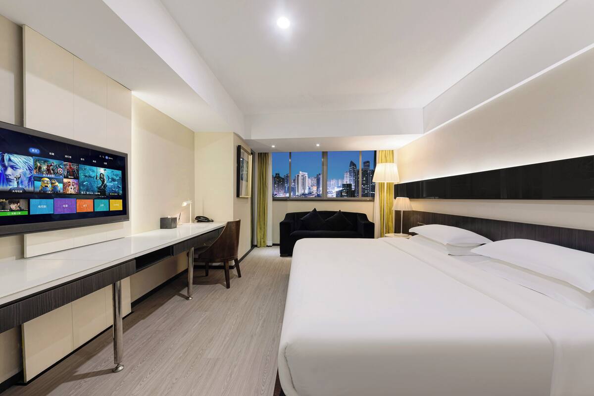A hotel room with a large flat-screen TV on a white desk, a bed with white linens, a black chair, and a view of the city skyline through the window.