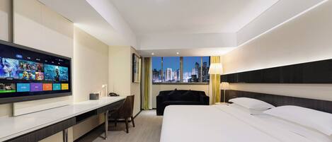 Habitación de hotel con una amplia televisión de pantalla plana sobre un escritorio blanco, cama con ropa de cama blanca, una silla negra y vista al horizonte de la ciudad a través de la ventana.