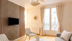 A modern living room with a flat-screen TV, two armchairs, a sofa, a round coffee table, and a large window with curtains. - Family flat -  Montparnasse (Paris)