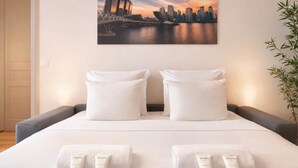 A hotel room with a bed, two white pillows, a nightstand with toiletries, and a cityscape painting on the wall. - Family flat -  Montparnasse (Paris)