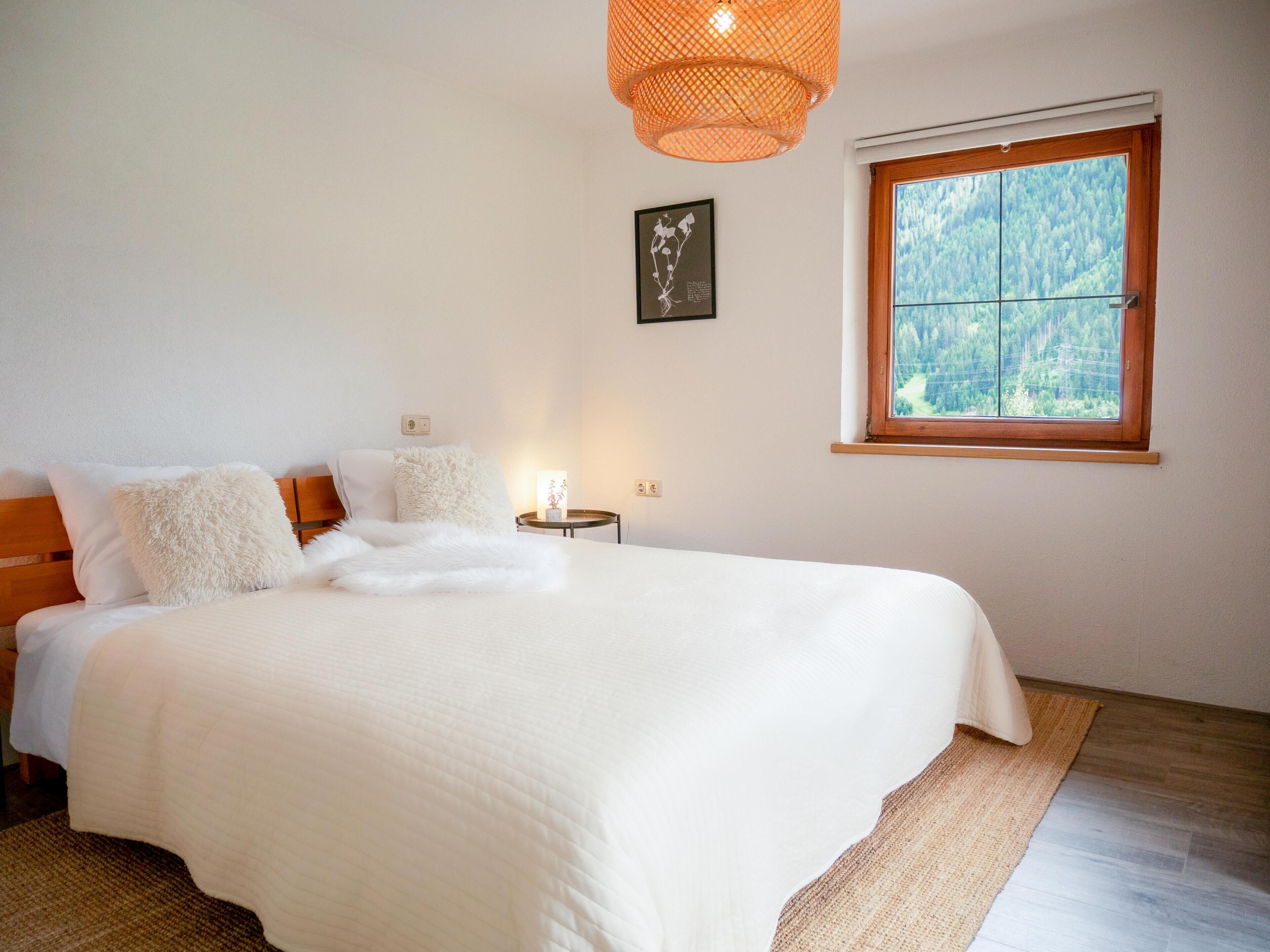 Ein Schlafzimmer mit einem großen Bett, einem Nachttisch mit Lampe, einem Fenster mit Blick auf Bäume und einem Wandbild.