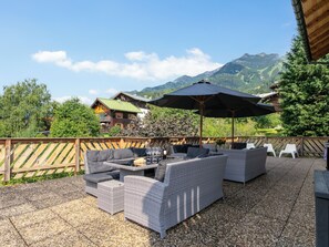 Eine Terrasss mit Korbmöbeln, einem großen Sonnenschirm und Blick auf Berge sowie Gebäude im Hintergrund.