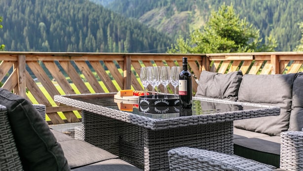 Un área de estar al aire libre con muebles de mimbre, una mesa, copas de vino y botellas, con vista a un paisaje montañoso.
