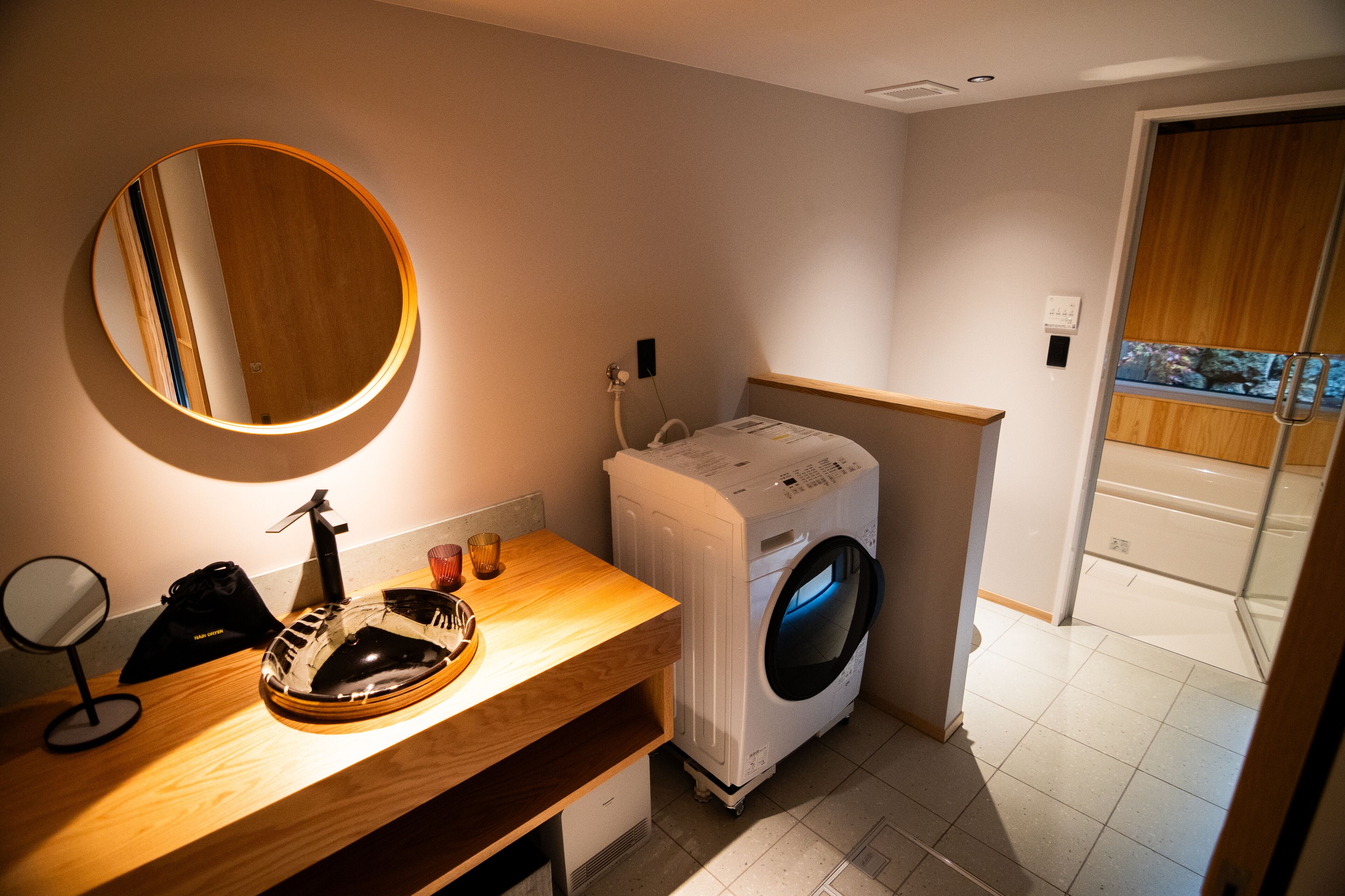 Une petite salle de bain avec un meuble-lavabo en bois, un miroir rond, un lave-linge et une vue sur un balcon à travers une porte vitrée.