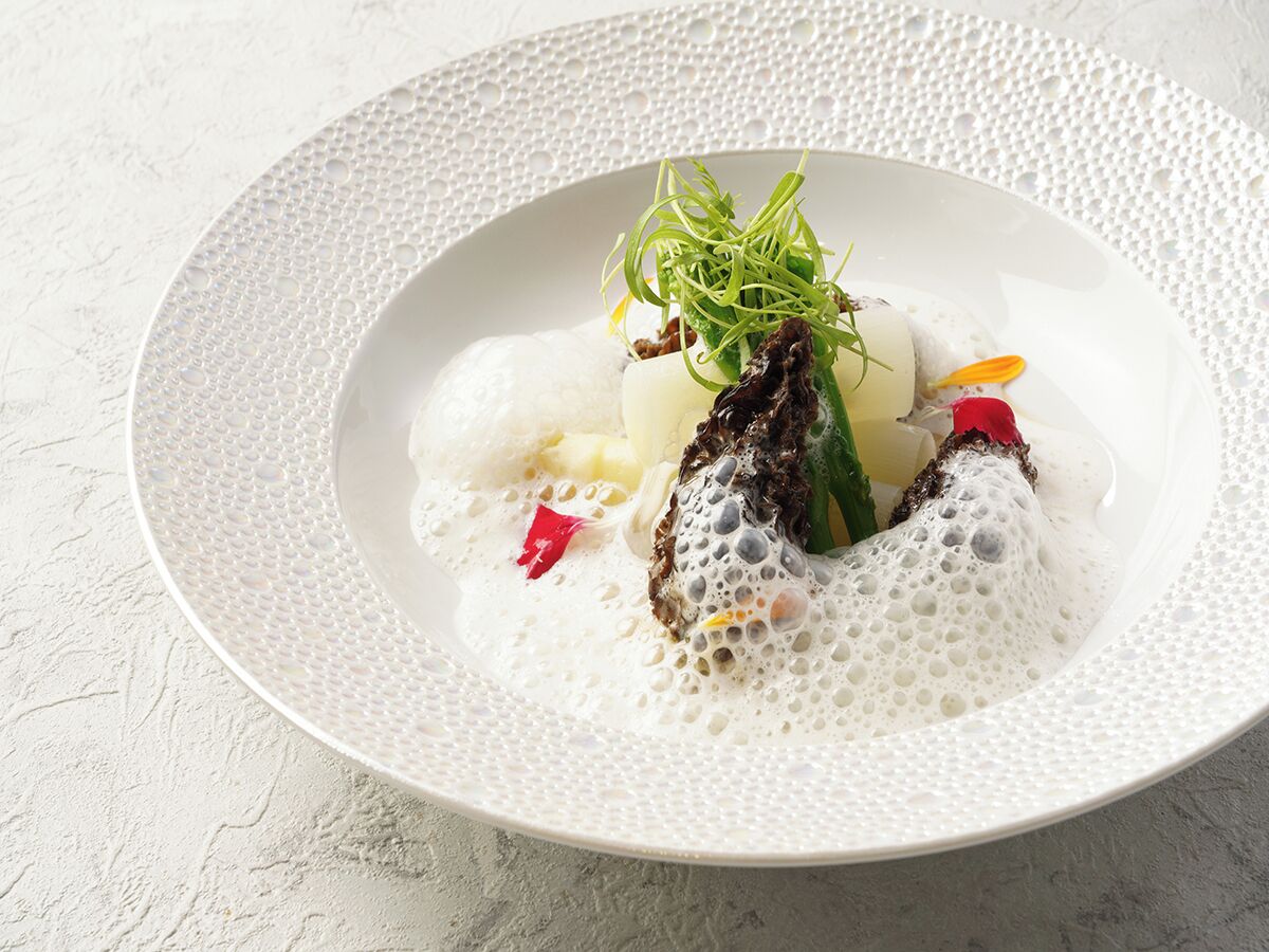 A white plate with a textured edge, containing a dish with white and dark elements, garnished with green and red accents.