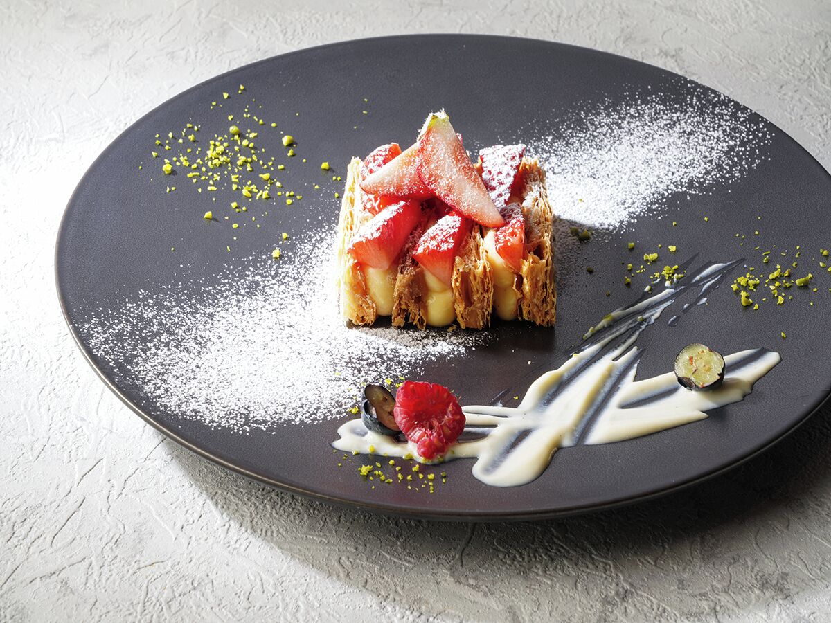 Un postre en un plato con azúcar glas, fruta y un hilo de salsa.
