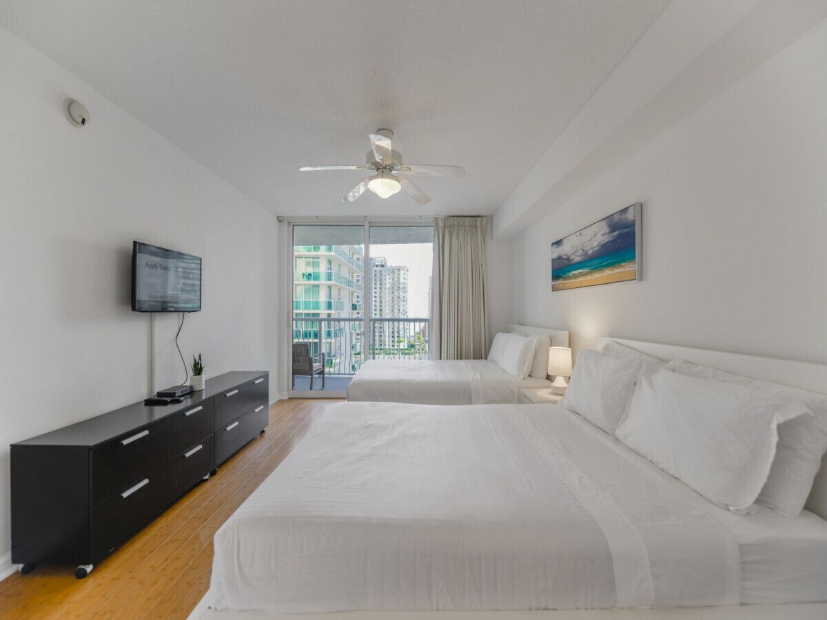 Een hotelkamer met een bed, een televisie, een bureau en een balkon met uitzicht op gebouwen.