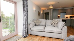 Ein modernes Wohnzimmer mit grauem Sofa, gemusterten Kissen, Holzboden und einer Schiebetür aus Glas. Im Hintergrund ist eine Küche zu sehen.