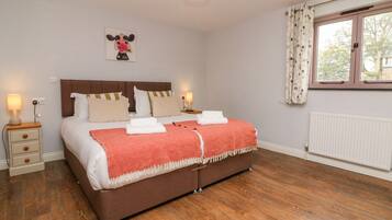 A bedroom with a wooden floor, a bed with a brown headboard, white bedding, and a red blanket. There is a window with curtains and a wall-mounted picture.